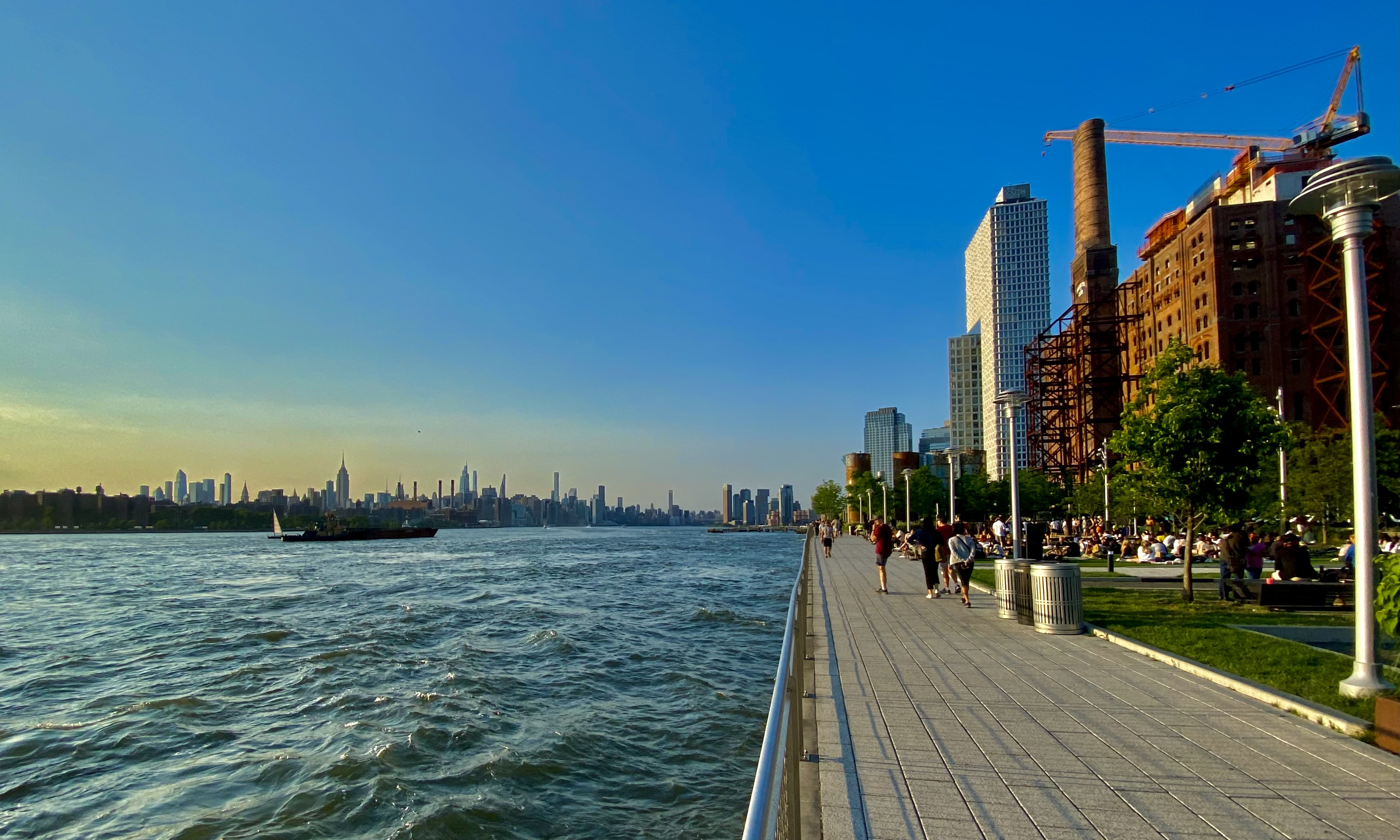 People stroll along Domino Park’s tree-lined waterfront beside the East River and the historic sugar refinery.